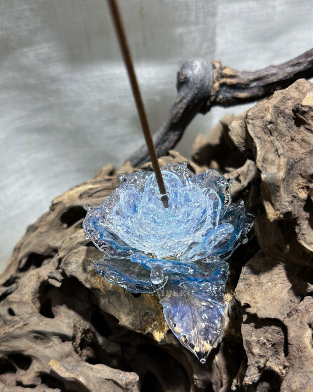 Light blue handcrafted lampwork glass rose incense holder by Ambero Oasis, resting on a piece of natural wood.