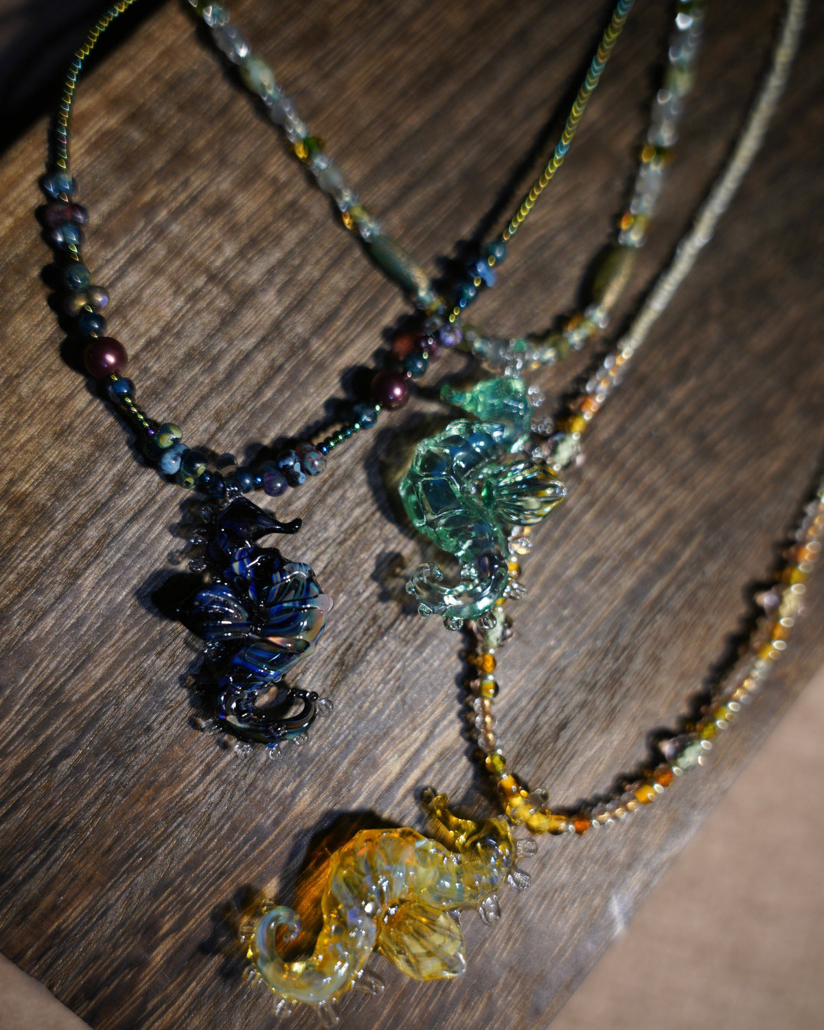 handmade lampwork glass seahorse pendant necklaces in blue, teal, and amber gold on wood background. Includes free titanium steel chain, beaded cord sold separately.