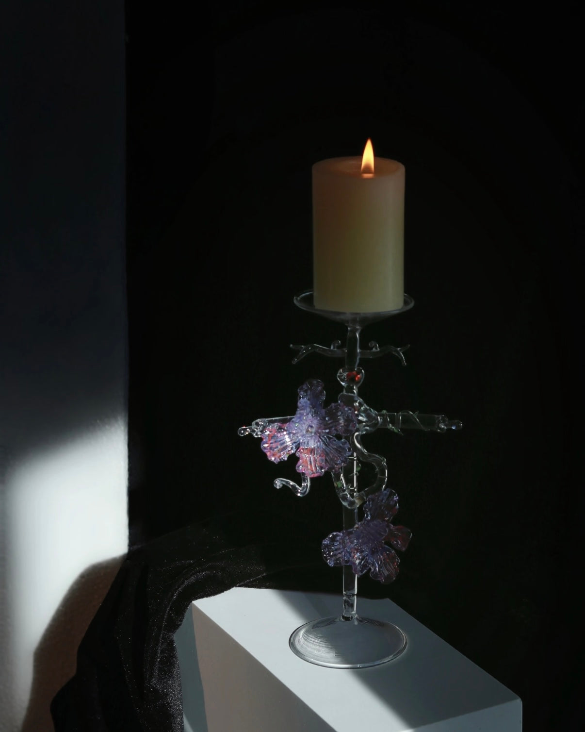 Purple lampworked glass cross iris candle holder on a light wooden table, with a lit taper candle glowing through the purple glass