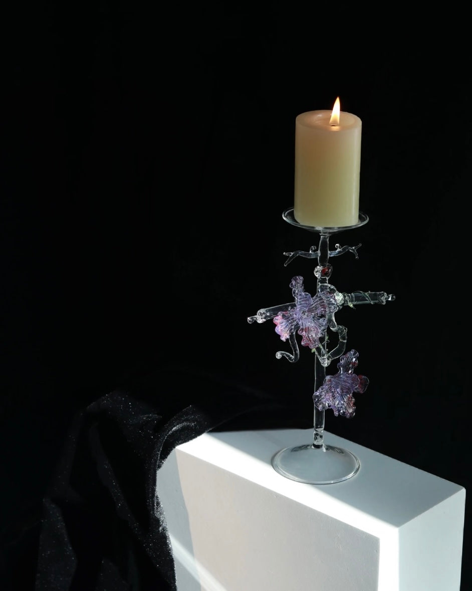 Purple lampworked glass cross iris candle holder on a light wooden table, with a lit taper candle glowing through the purple glass