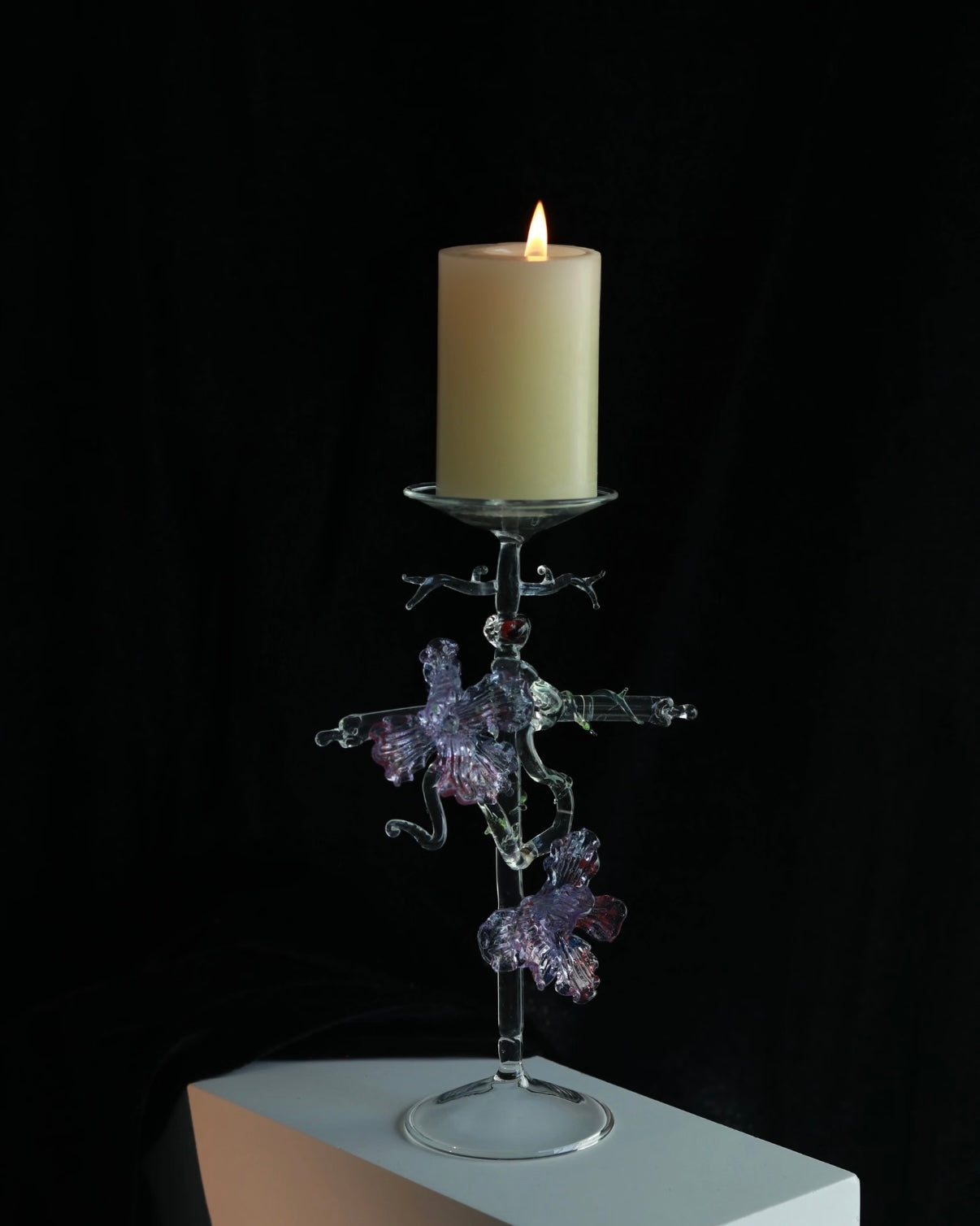 Purple lampworked glass cross iris candle holder on a light wooden table, with a lit taper candle glowing through the purple glass