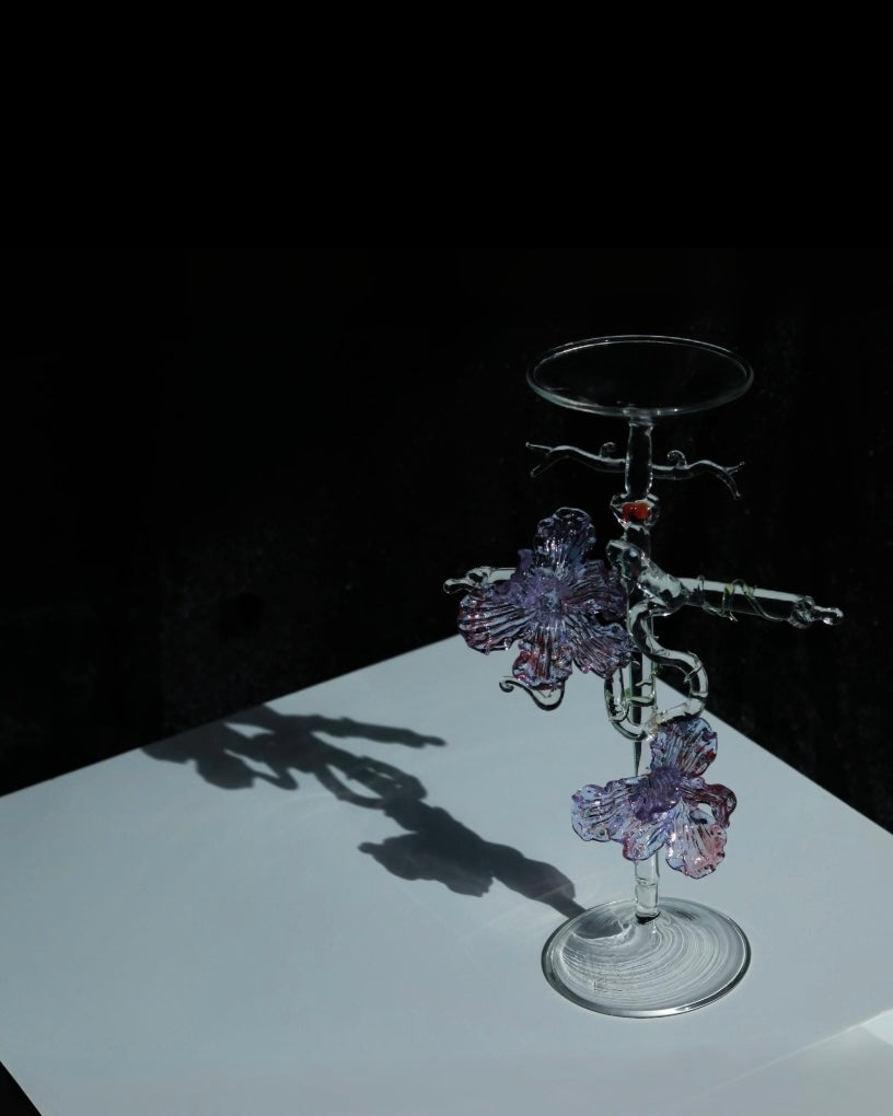 Purple lampworked glass cross iris candle holder on a light wooden table, with a lit taper candle glowing through the purple glass