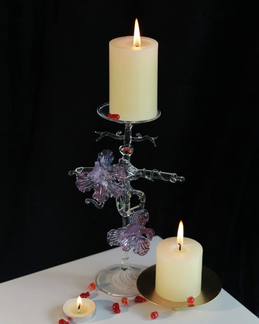 Purple lampworked glass cross iris candle holder on a light wooden table, with a lit taper candle glowing through the purple glass